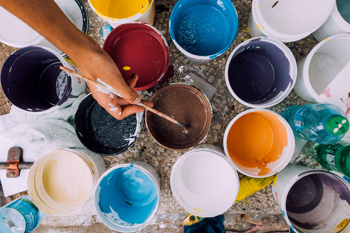 Artist holding paint brush and dipping it into buckets of colorful paint.