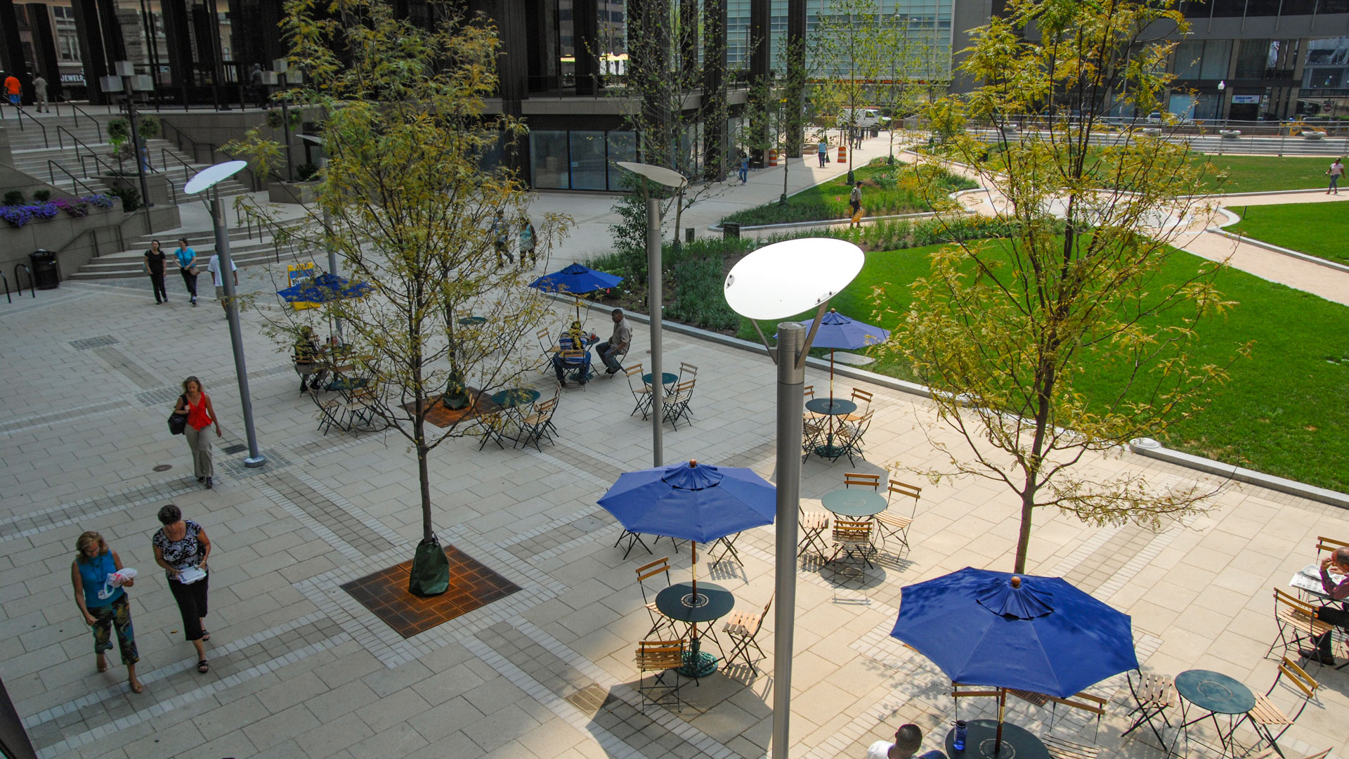 Overhead view of Downtown Baltimore's Center Plaza, featuring green grass, pathways, bistro tables, umbrellas, and walking pedestrians.
