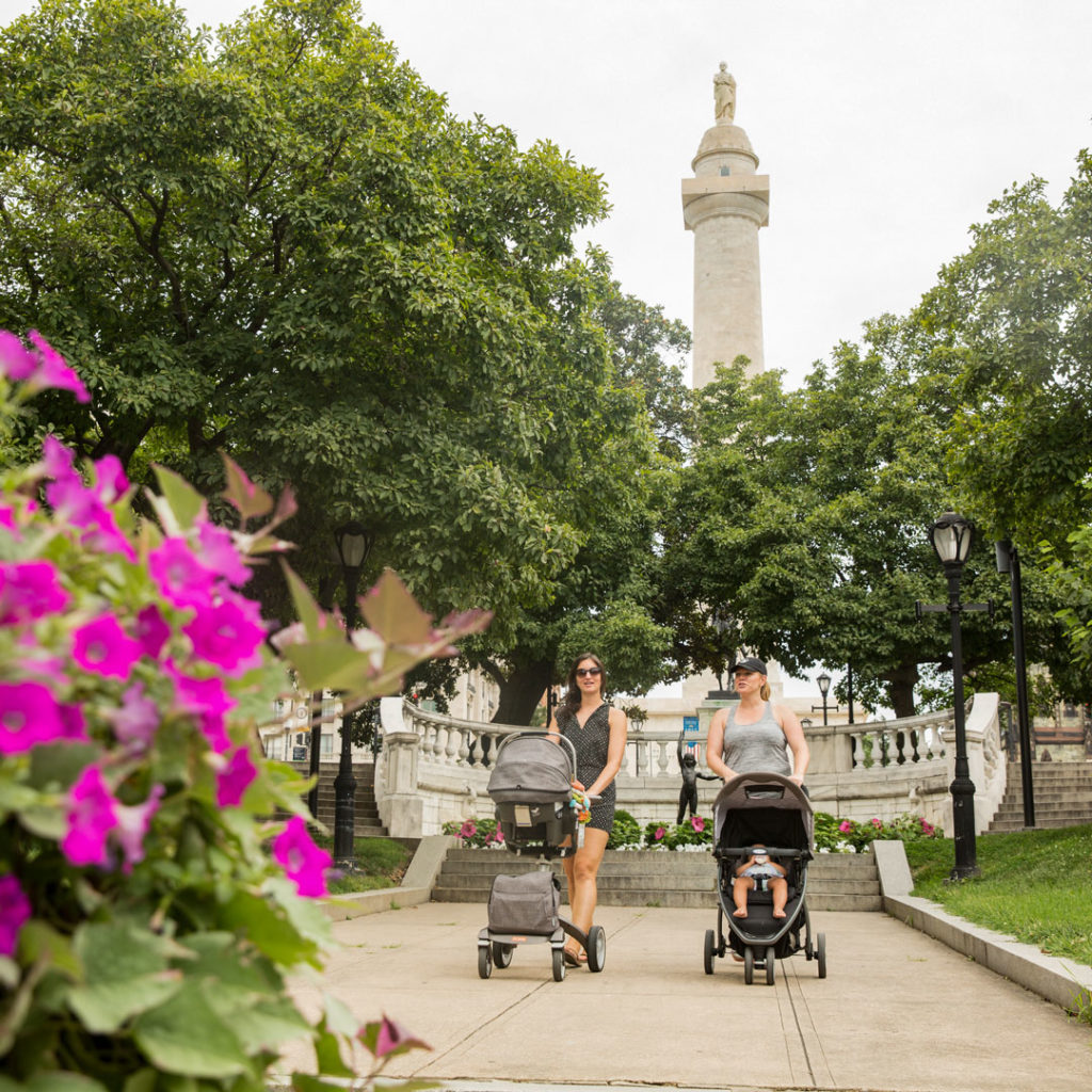 Two moms pushing strollers walking through Mt. Vernon Place park in Baltimore with the Washington Monument in the background.