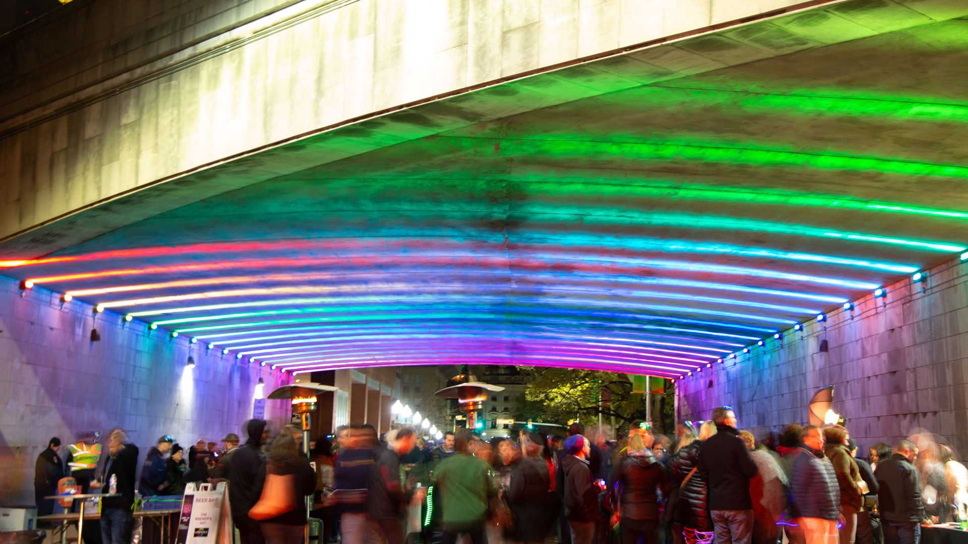 Crowd under colorful viaduct lights for Downtown Partnership's Lit City event.