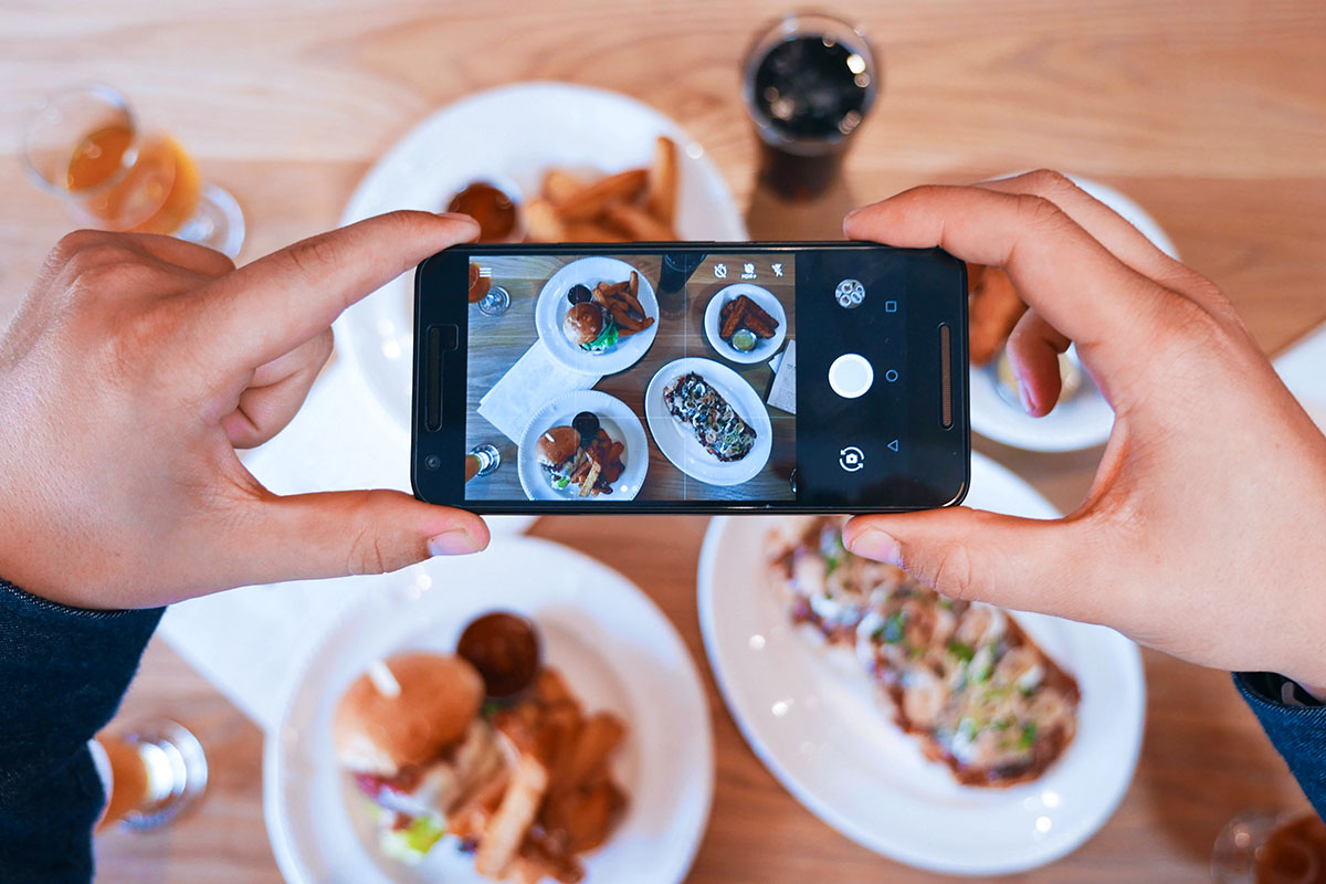 Hands holding cell phone, taking photo of meal on tabletop.