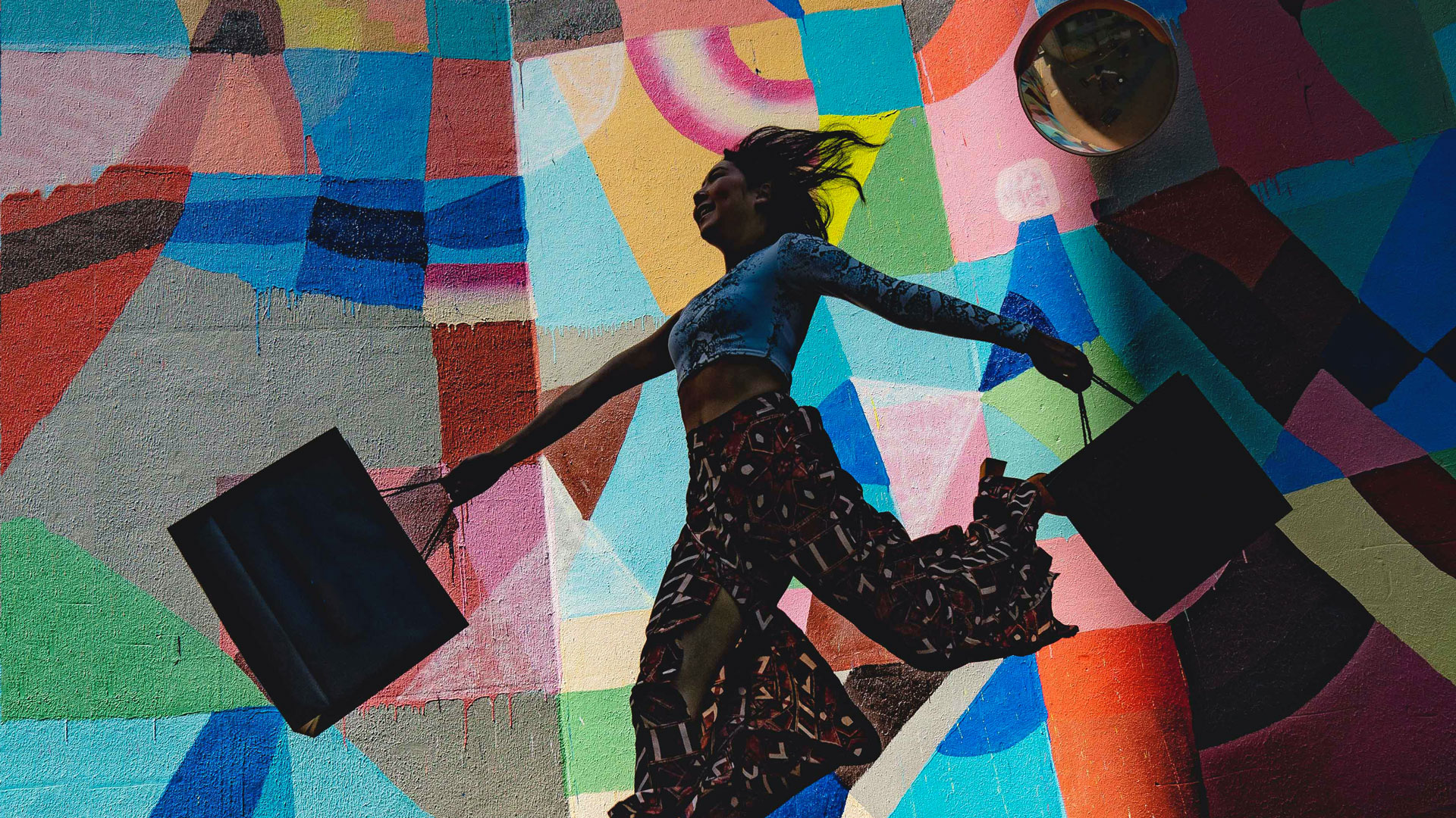 woman skipping and holding two shopping bags in front of colorful, abstract mural.